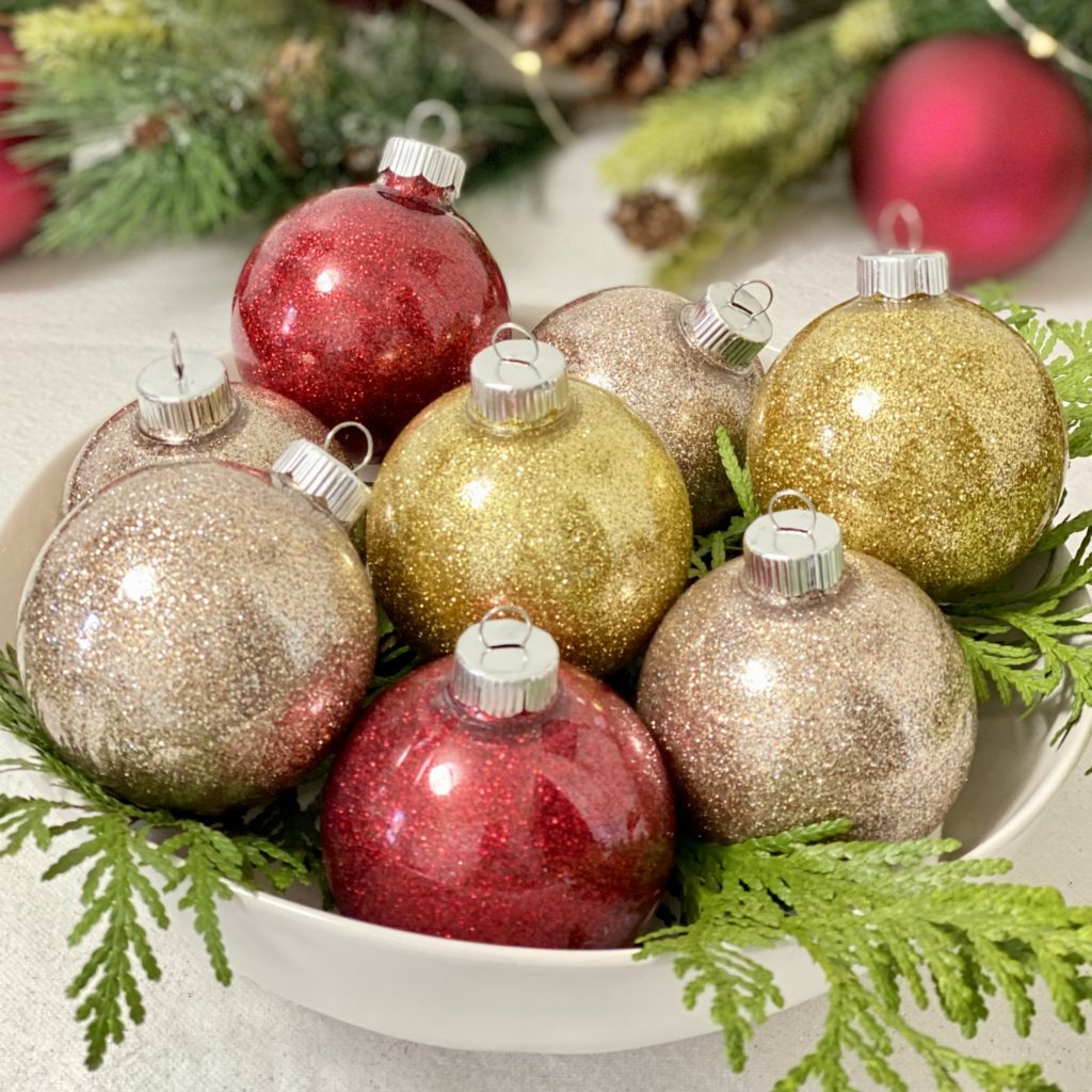 Red, gold, and champagne colored glitter ornaments in a bowl with evergreens.