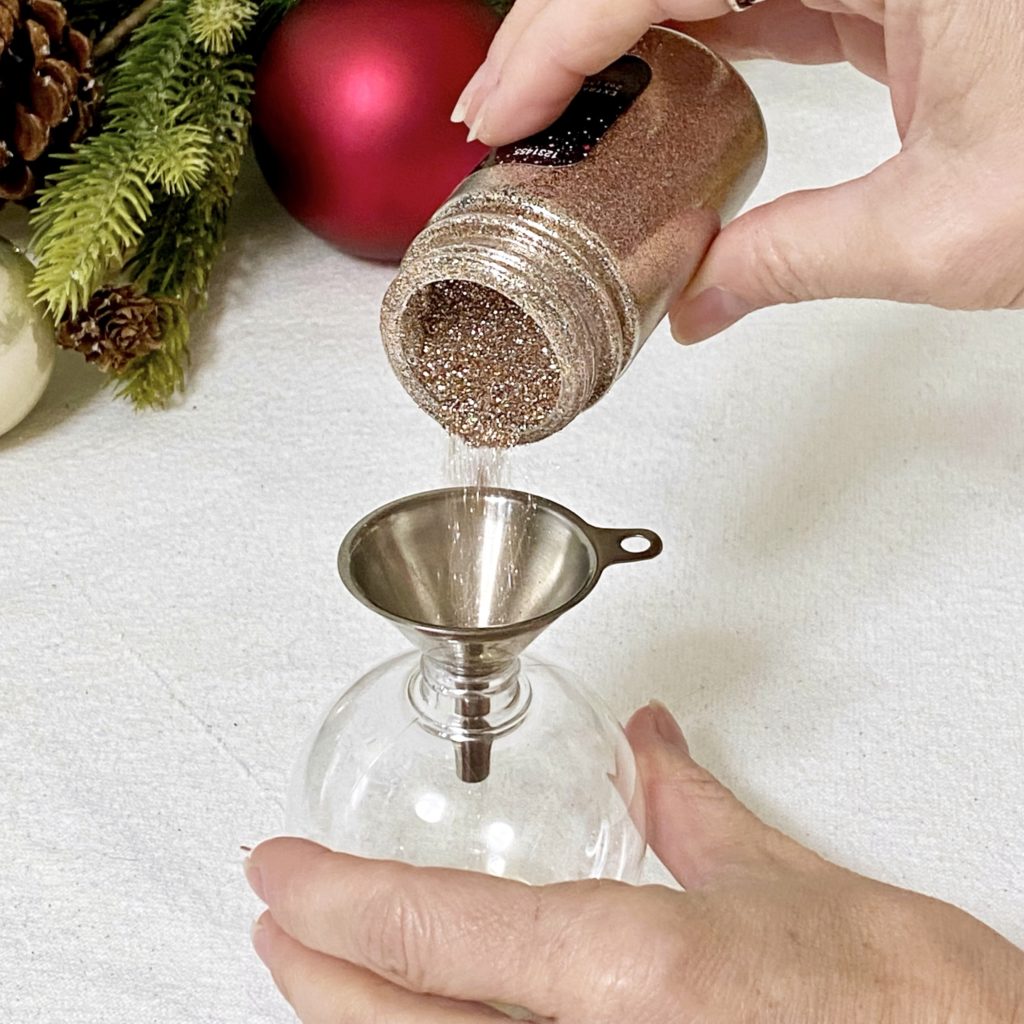 Pouring extra fine glitter into a clear plastic ornament through a funnel.
