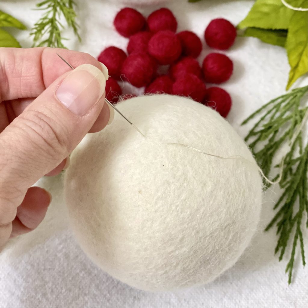 Pulling a needle and thread through a white wool dryer ball to attach a hanging loop on the ornament.