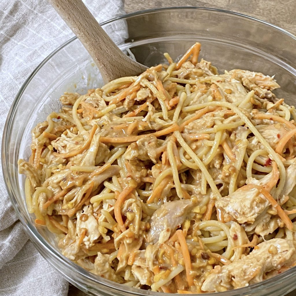 Chicken and noodles with peanut sauce being stirred in a glass bowl.
