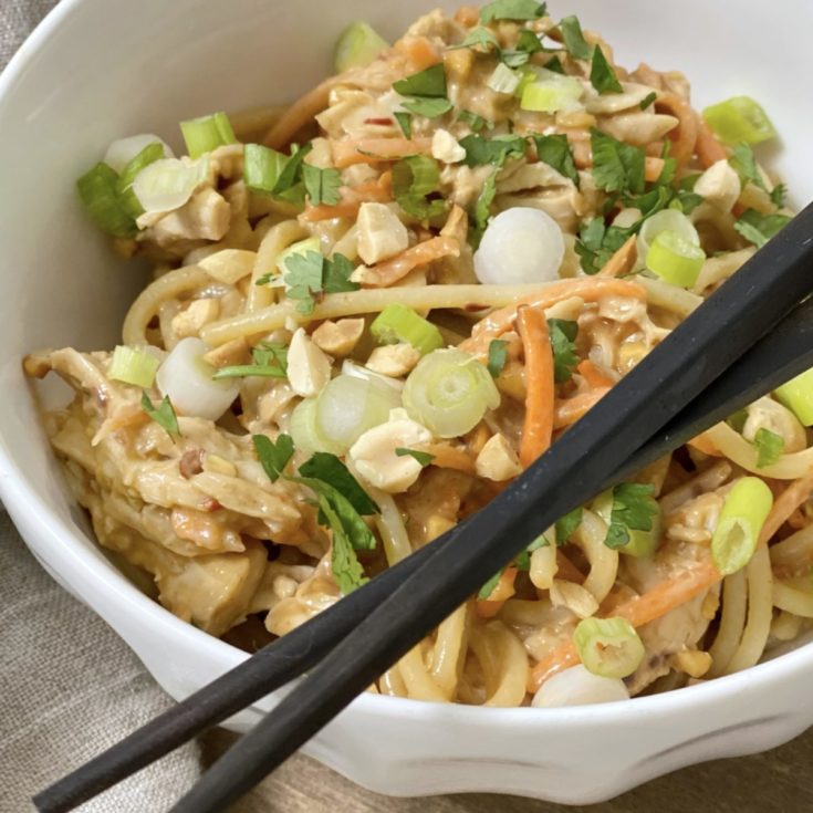 A close up photo of chicken and noodles with peanut sauce in a white bowl with chopsticks.