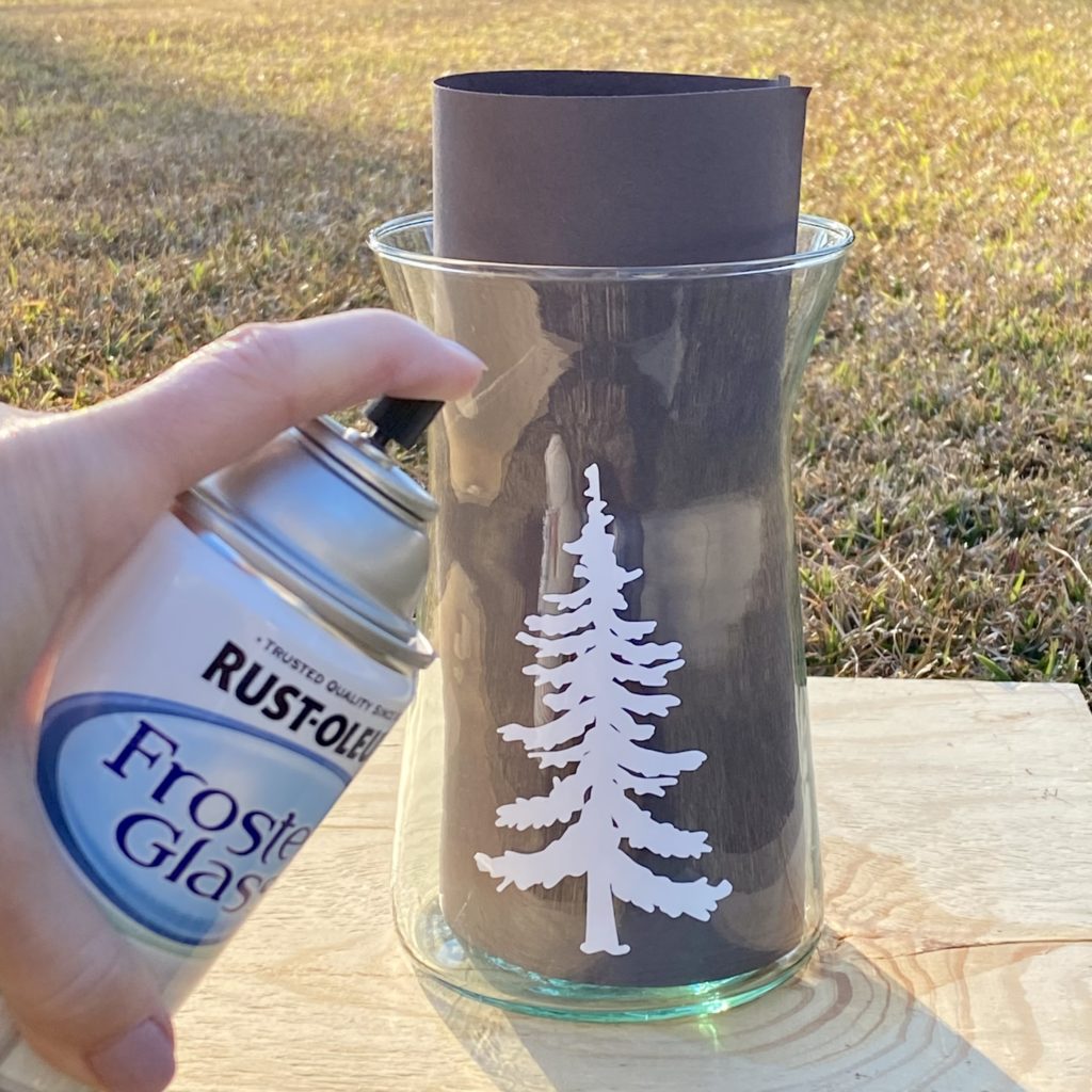 Spraying a glass vessel with an evergreen tree decal on it with frosted glass spray paint outside.