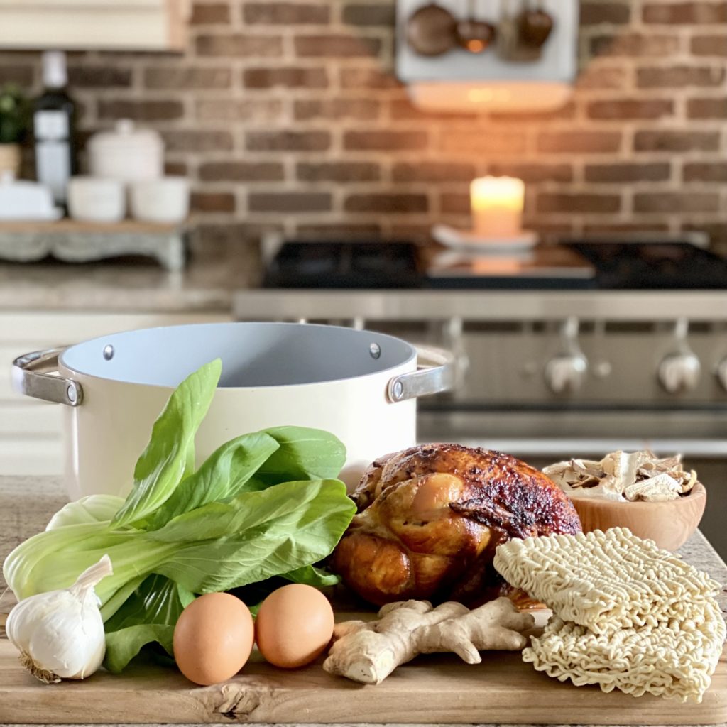 Some the ingredients in easy weeknight chicken ramen including rotisserie chicken, ramen noodles, fresh bok choy and more set out on the kitchen island in front of the stove with a soup pot.