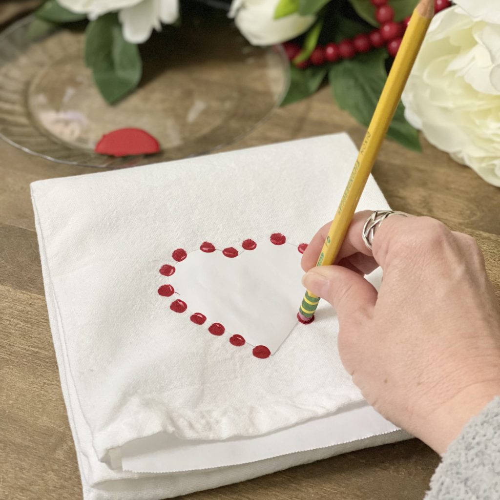 Stamping red fabric paint around the freezer paper heart on the dish towel with a pencil.