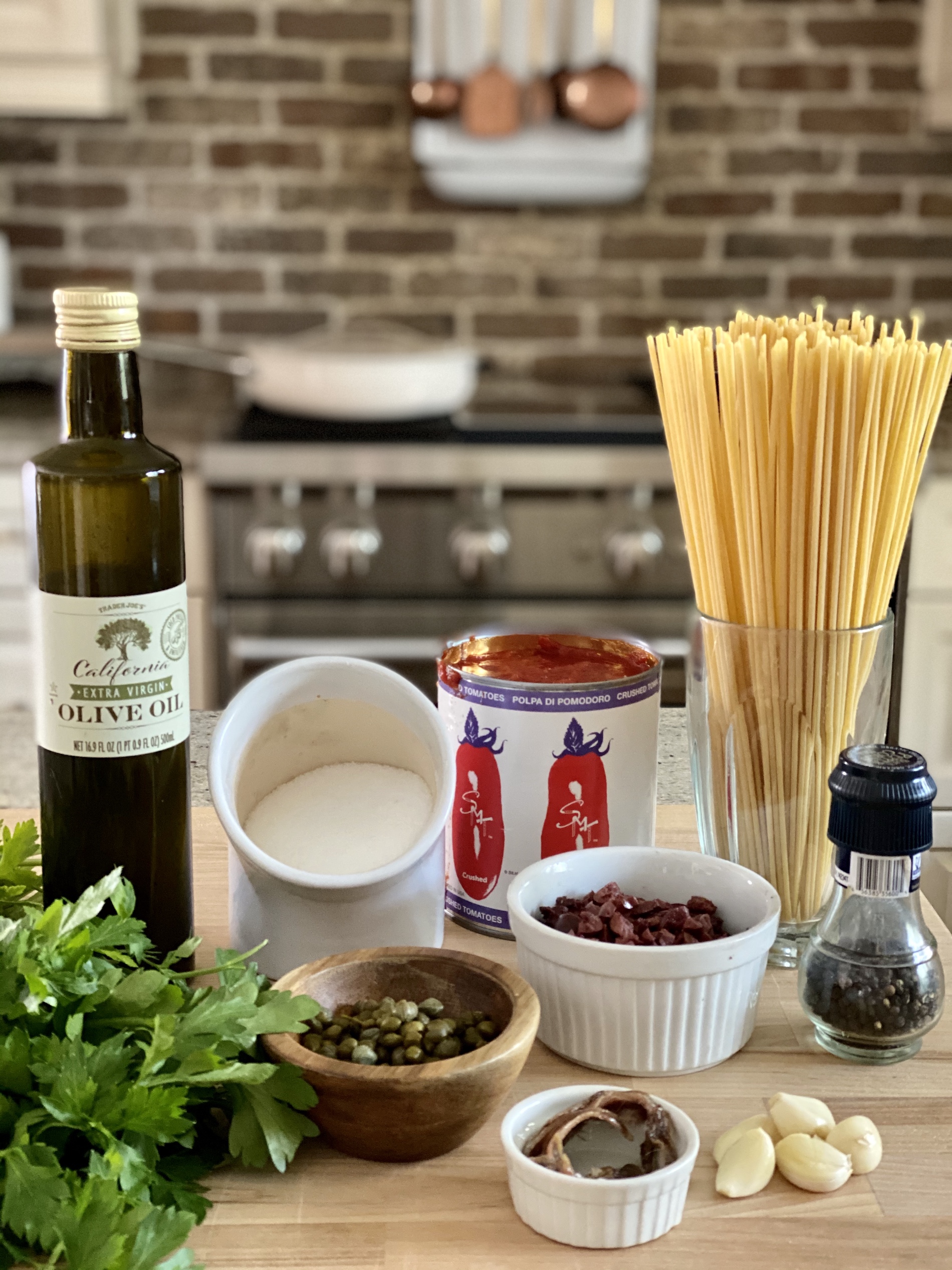 Pasta Puttanesca ingredients including olive oil. salt, pepper, tomatoes, pasta, kalamata olives, capers, red pepper flakes, anchovies, and parsley