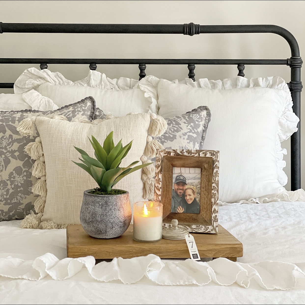 Bedside essentials: faux plant, candle, and picture frame arranged on a tray on the bed.