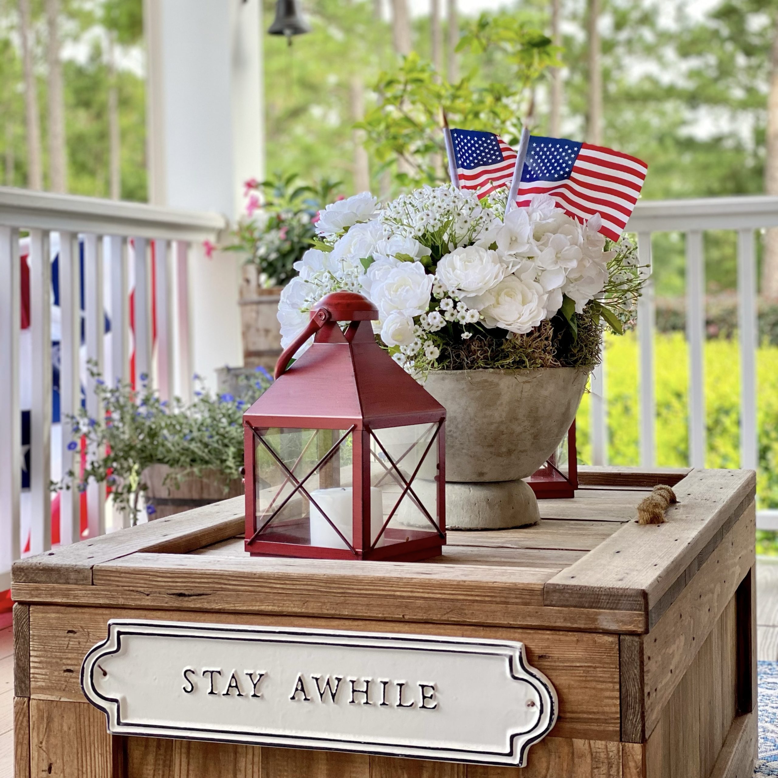 Wood trunk coffee table styled with a red lantern and a white flower arrangement in a concrete pedestal bowl with American flags.