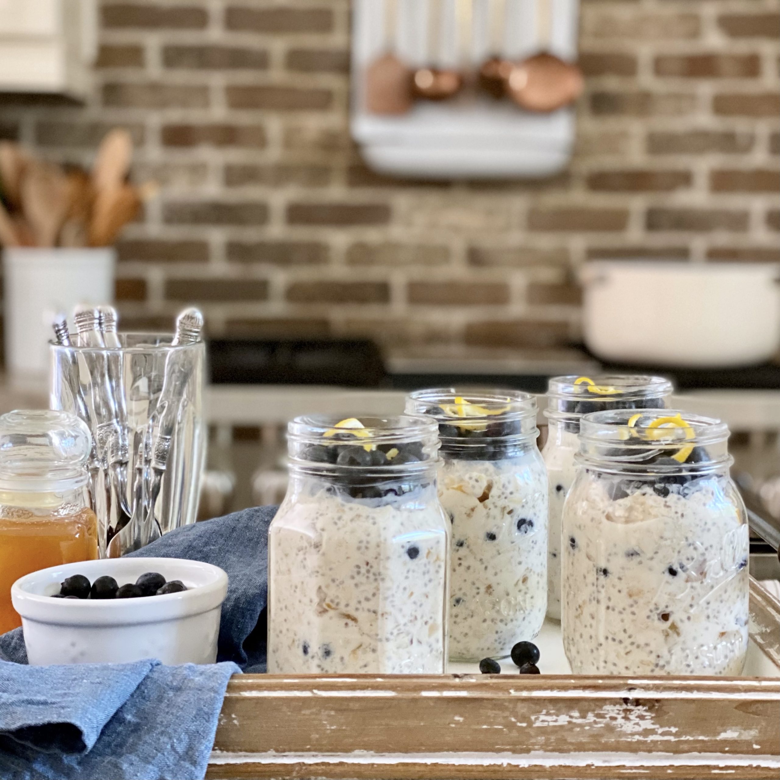 Four jars of overnight oats with blueberries and lemond zest on top on a tray in the kitchen with spoons , extra blueberries in a dish, and honey.