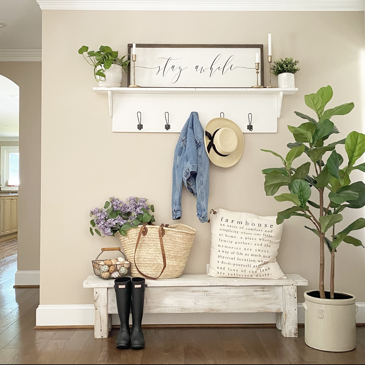 Farmhouse foyer styled with a white wood shelf and hooks. There is a farmhouse sign on self that says, "Stay awhile." A denim jacket and straw hat hang from the hooks.