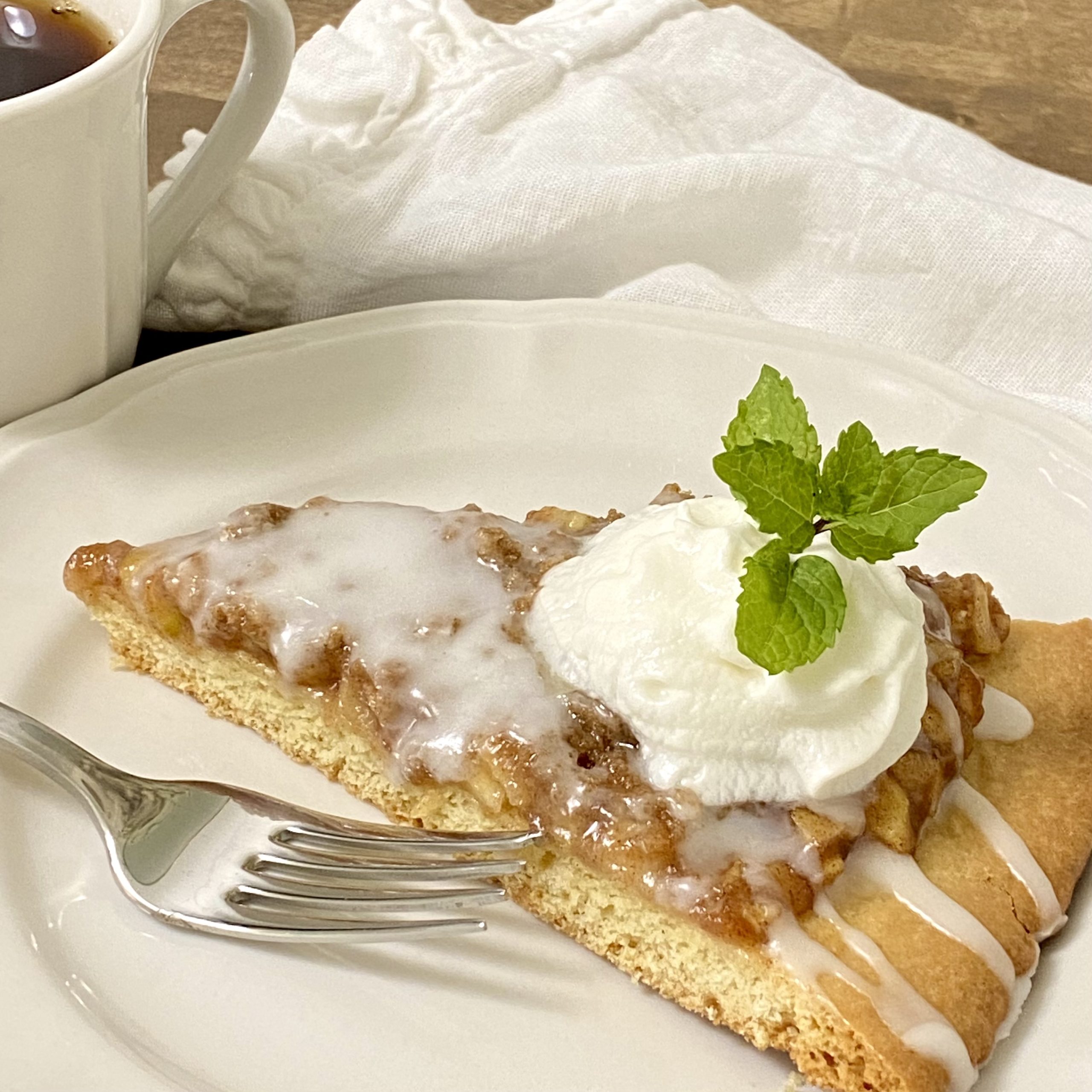 A slice of apple pie pizza with a dollop of whipped cream and a mint sprig on a white a white plate.