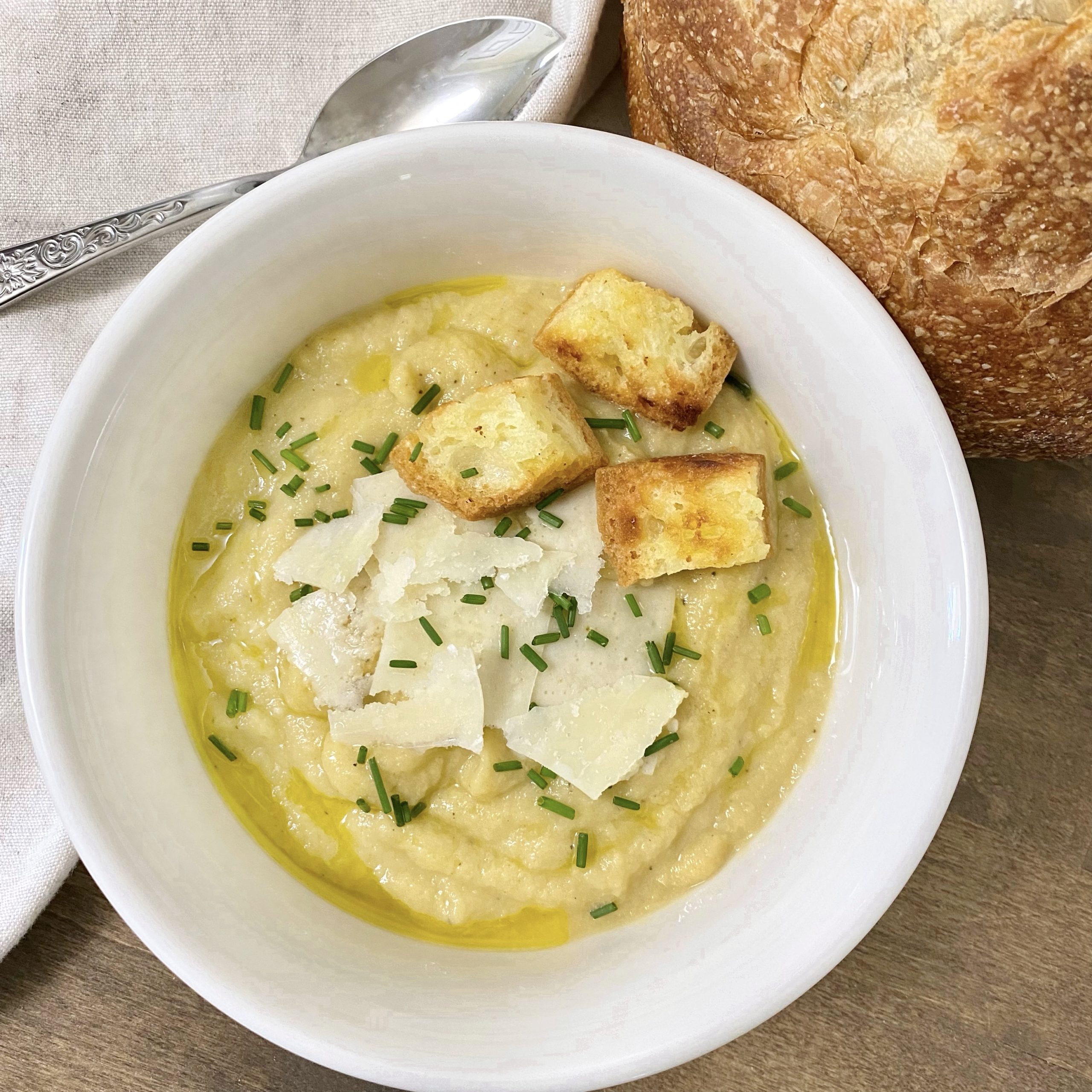 Creamy cauliflower soup with Basil Olive Oil in a white bowl garnished with parmesan cheese, chives, and croutons with a spoon and rustic bread on the side.