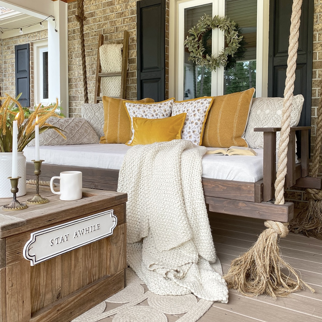 Farmhouse front porch swing with muted gold and cream throw pillows and a blanket on it. In front of the swing is a wood chest coffee table with a mug, brass candlesticks and a crock with fall stems in it.