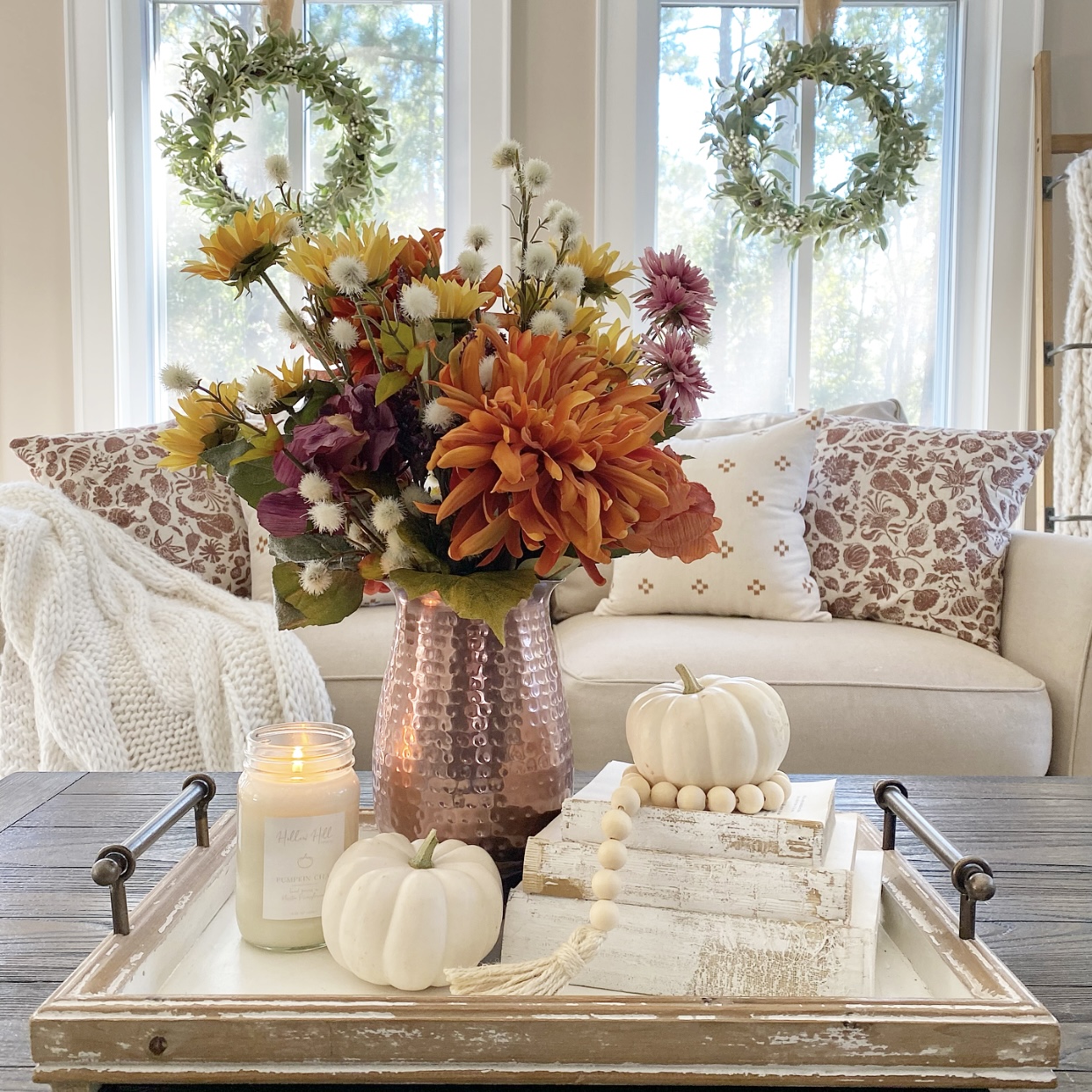 A Fall vignette with flowers in a copper pitcher, vintage books, pumpkins, wood beads, and a candle on a tray on the coffee table in front of the sofa.