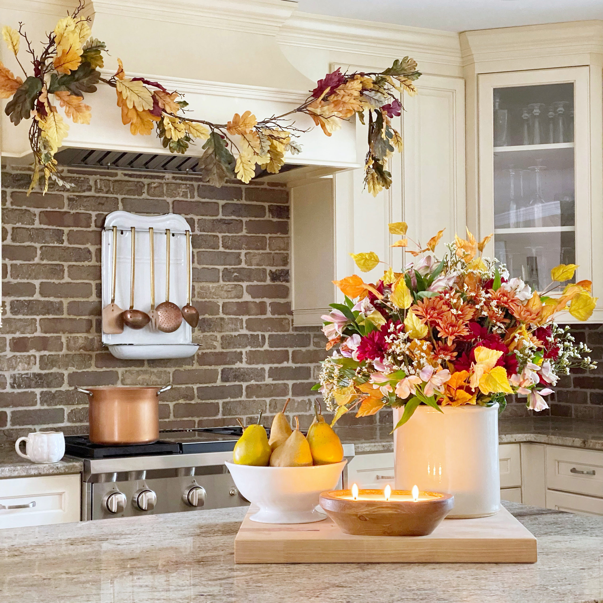 Kitchen island with a cutting board on it styled with a fall floral arrangement, a bowl of pears, and a candle. In the background is the range an on the hood is a vibrant fall colored garland.
