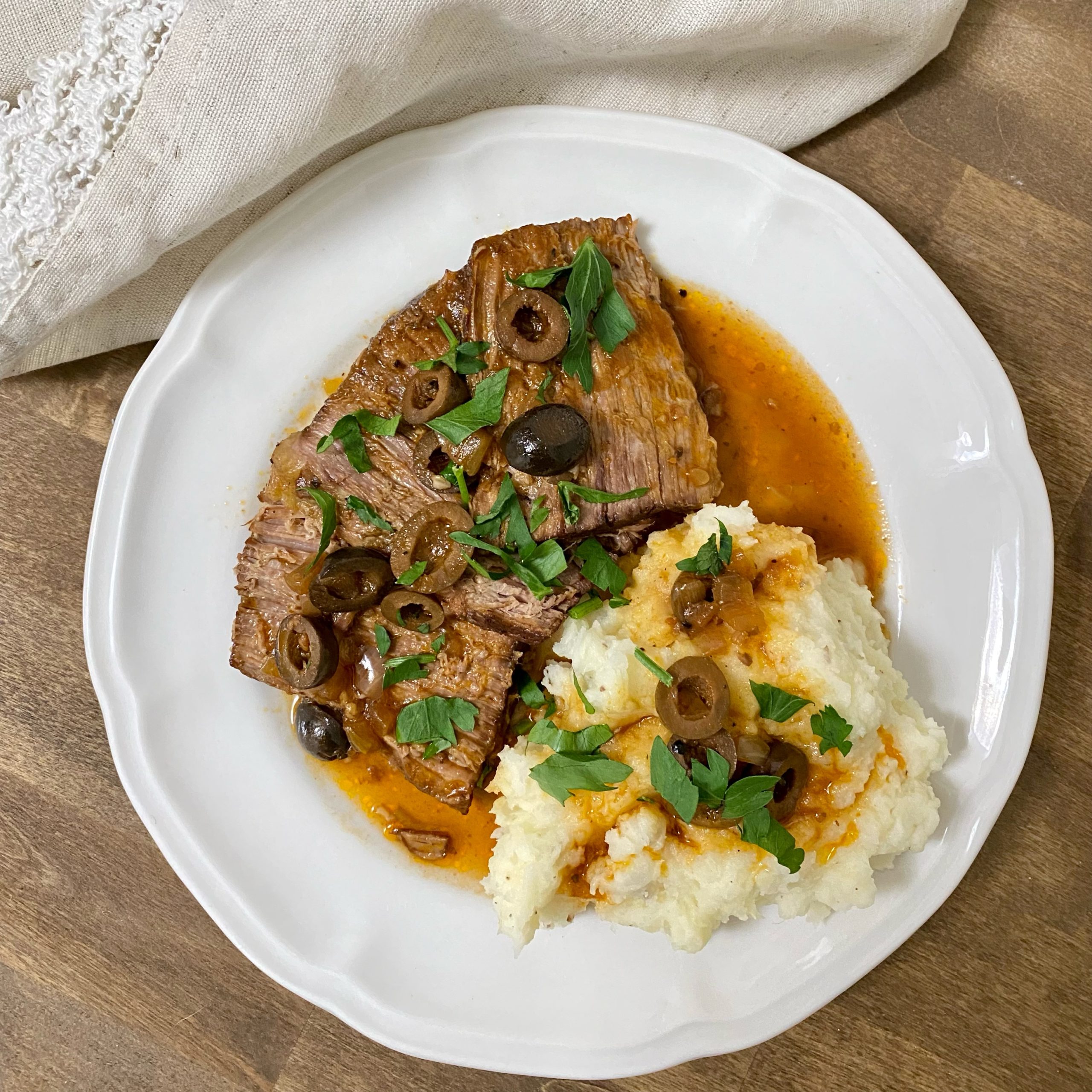 Italian pot roast on a white plate with mashed potatoes and gravey garnished with fresh chopped parsley.