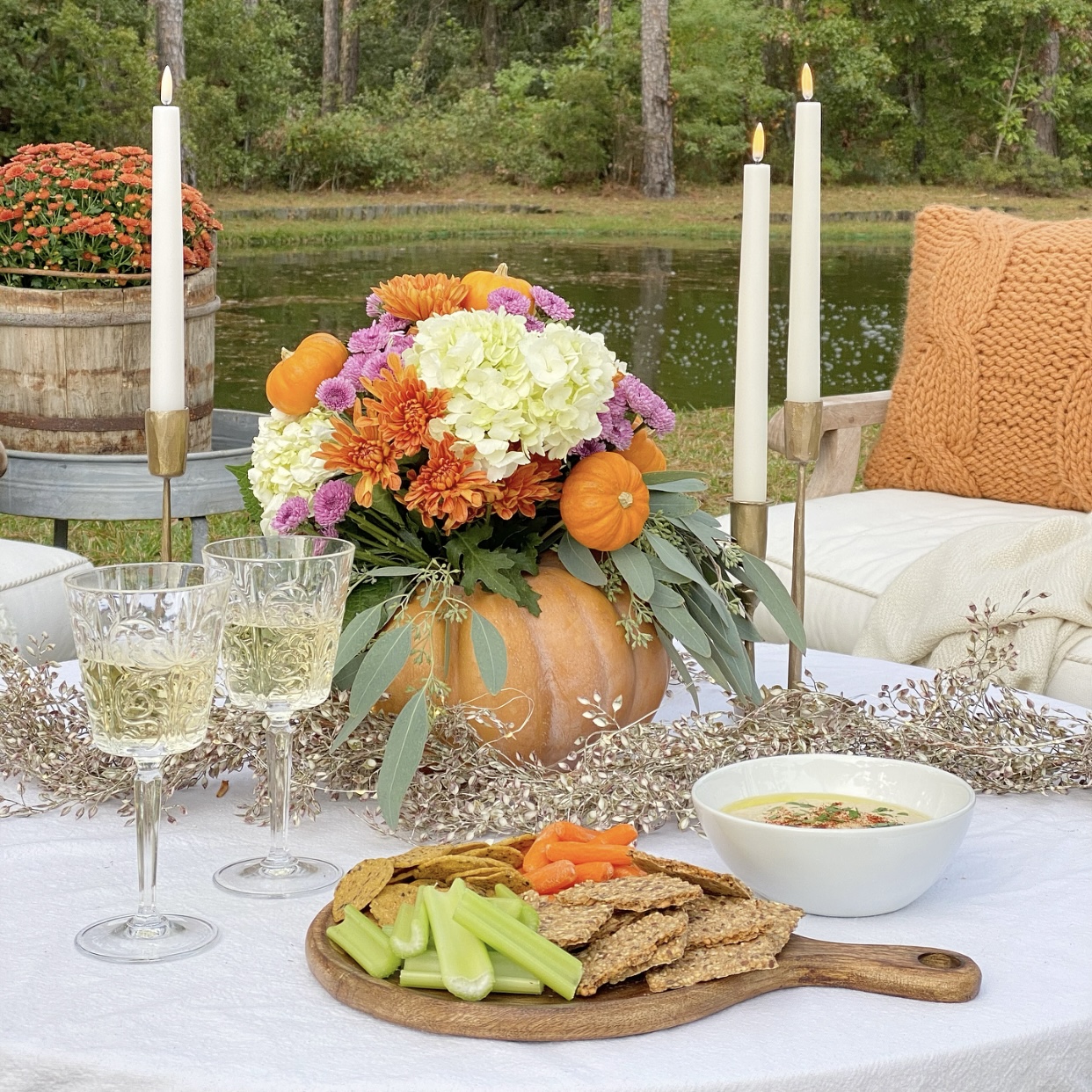 Fall floral arrangement in a hallowed out pumpkin, candles, wine, and white bean hummus on the tablea. In nthe background is a pond.