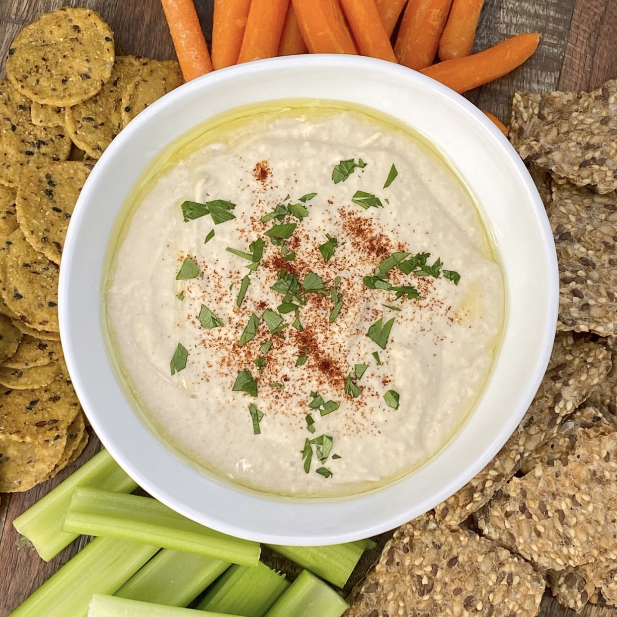 White bean hummus in a white bowl with crackers, celery sticks, and carrots around it. Perfect for a casual outdoor fall gathering.