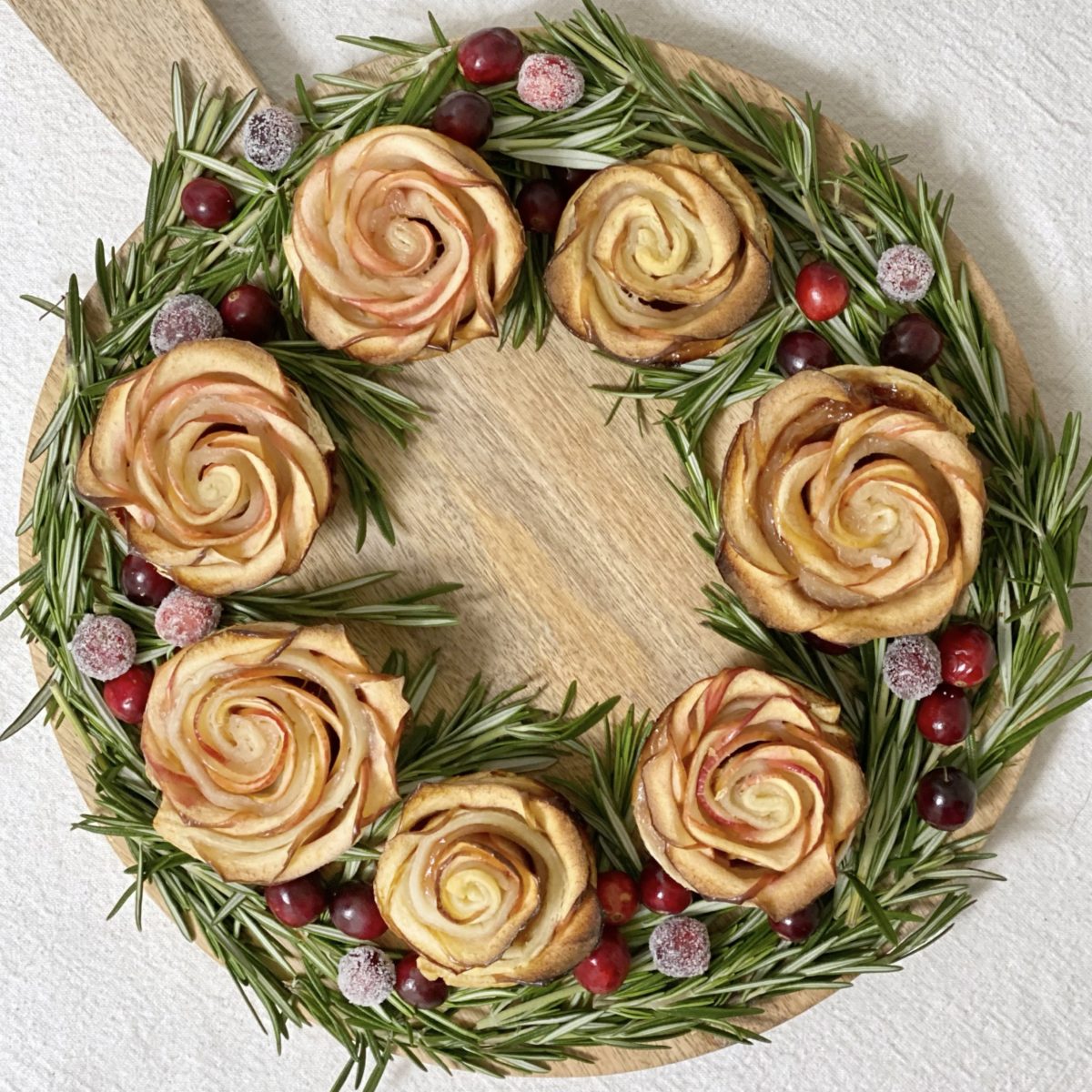 Baked apple roses on a round wood cutting board with rosemary and cranberries around them to make them look like a wreath.