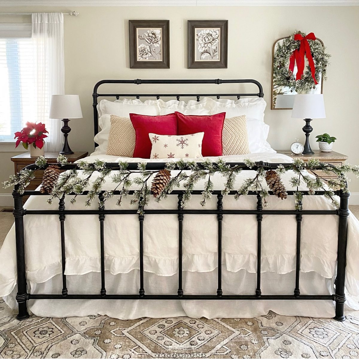 Iron bed with white linen bedding and red pillows. On the footboard it green garland and there is a wreath on the wall mirror over a nightstand.