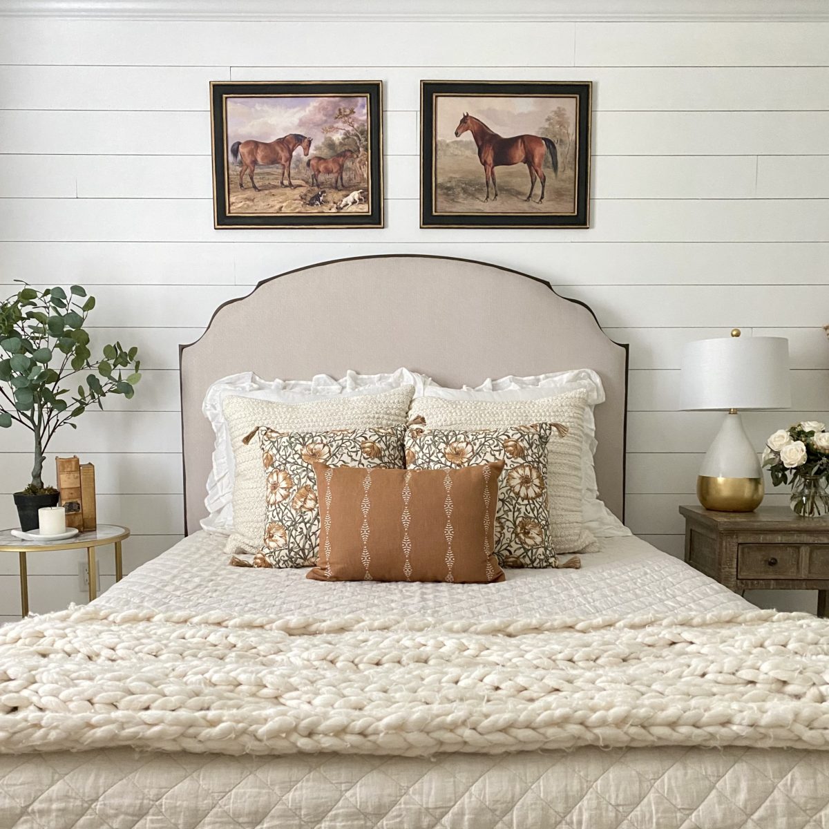 Eauropean farmhouse style Wesley Allen Chamberlain bed headboard with pillows on the bed below it. Above on the wall are horse prints and to the sides of the beds are nightstands with a lamp, flowers, and books on them.