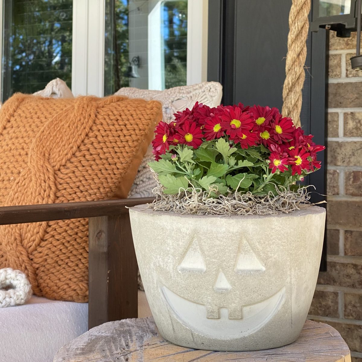 A concrete Jack O’Lantern planter with red mums in it on the porch.