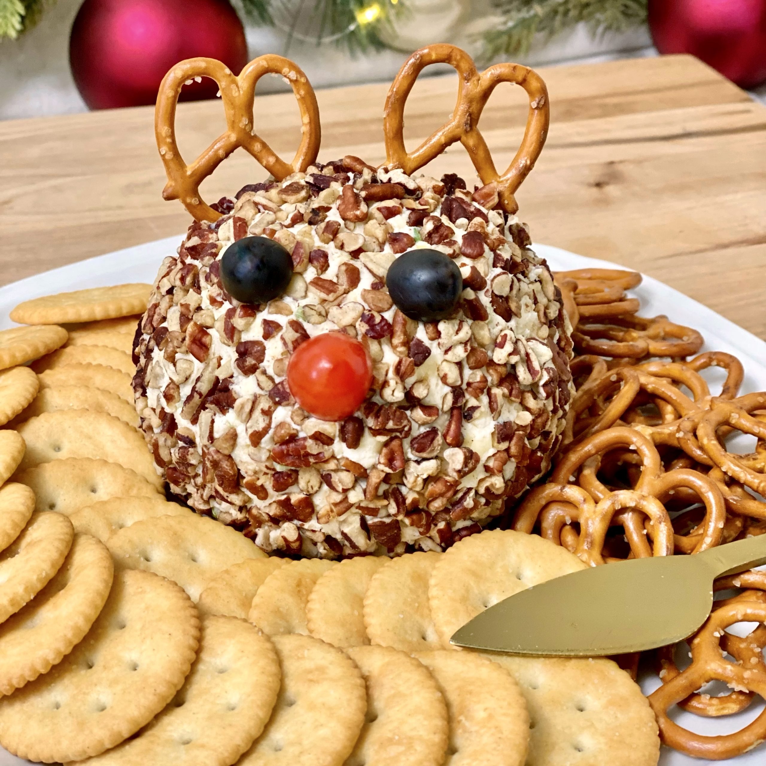 Rudolph Christmas Cheese Ball With crackers, pretzels, and a gold spreader with it on the plate.