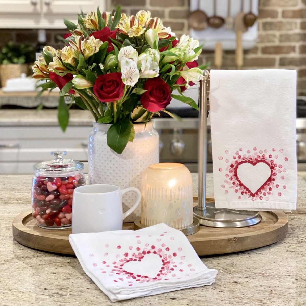 Beautiful and easy Valentine dish towels displayed on the island in the kitchen with some candy, a mug, flowers, and a candle on a tray.